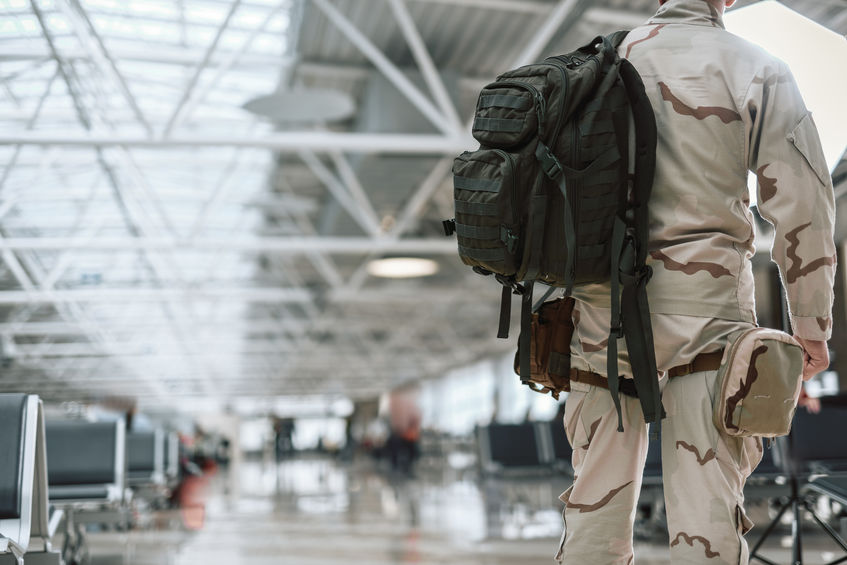 A soldier in camouflage uniform with a backpack stands in an empty airport terminal facing large windows.