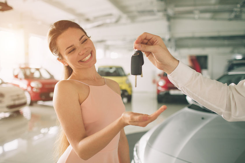 Person smiling and receiving a car key from another individual inside a car dealership showroom.