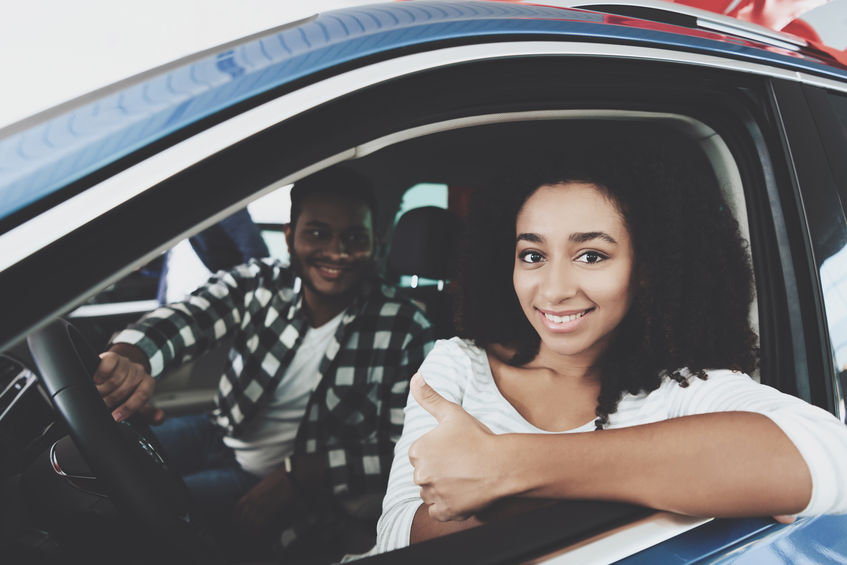 Woman in driver's seat of a car giving a thumbs up, with a man sitting in the passenger seat, both smiling.